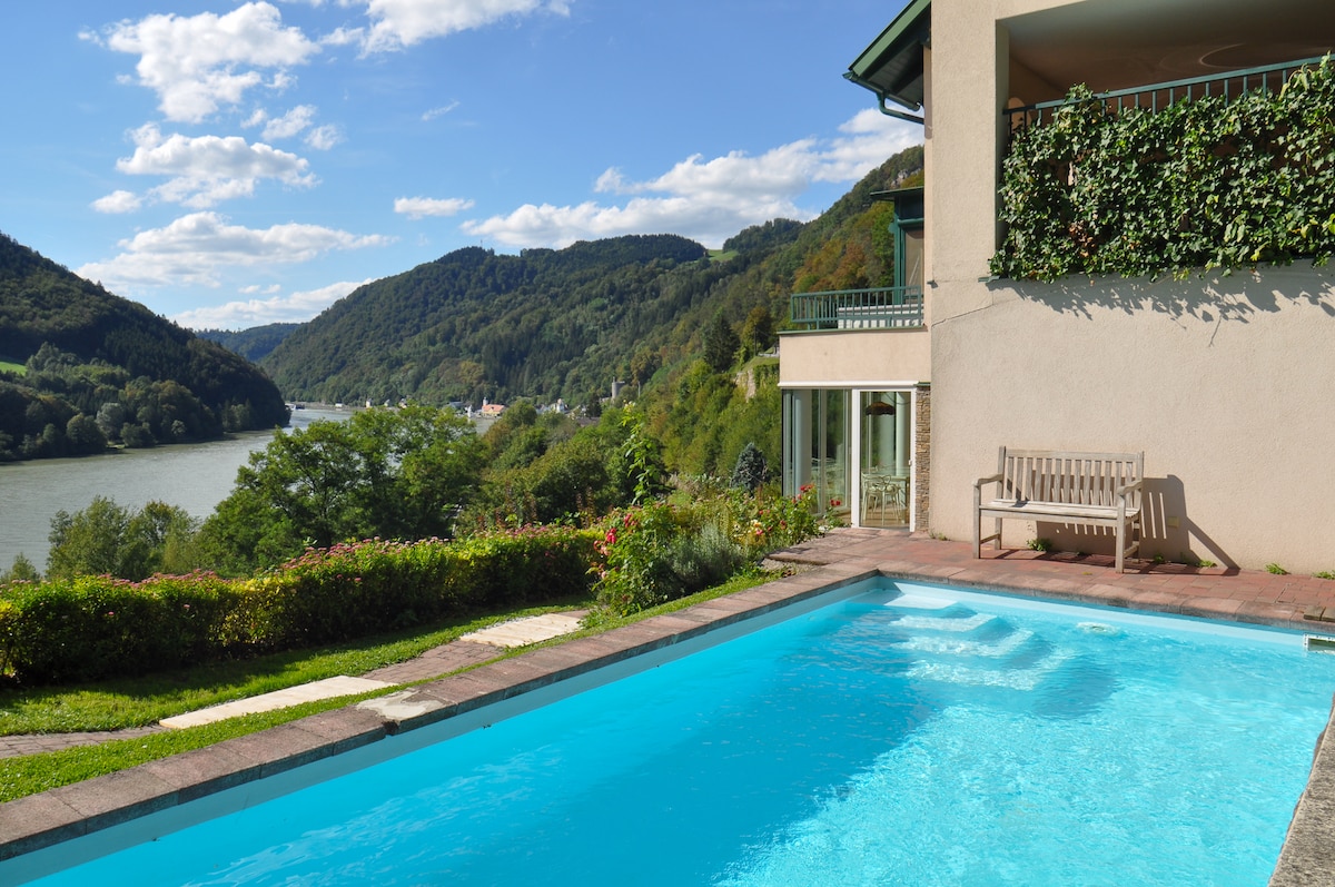 A serene outdoor space is showcased with a clear swimming pool reflecting the sky. Lush greenery surrounds the area, complemented by a wooden bench positioned nearby. The scenic view highlights the river and rolling hills in the background, enhancing the tranquil atmosphere.