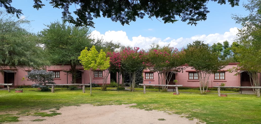 A series of single-story pink apartment buildings is set amid lush greenery. Flowering trees and well-maintained shrubs surround the area, with a grassy lawn leading to the entrance. Blue skies and scattered clouds are visible overhead, enhancing the inviting natural environment.