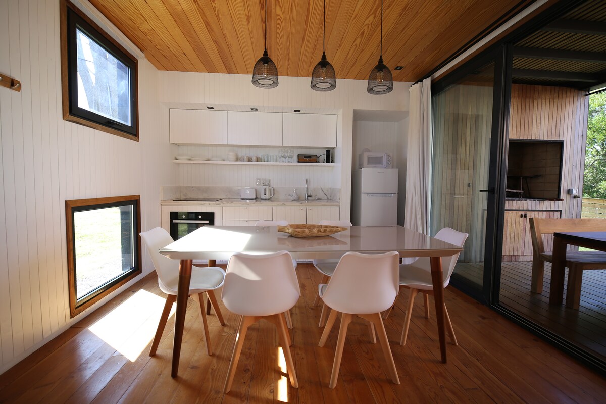 A bright dining area features a wooden table surrounded by six white chairs. Large windows bathe the space in natural light, while light wood paneling and an overhead light fixture contribute to a clean, contemporary aesthetic. A small kitchen area is visible in the background.