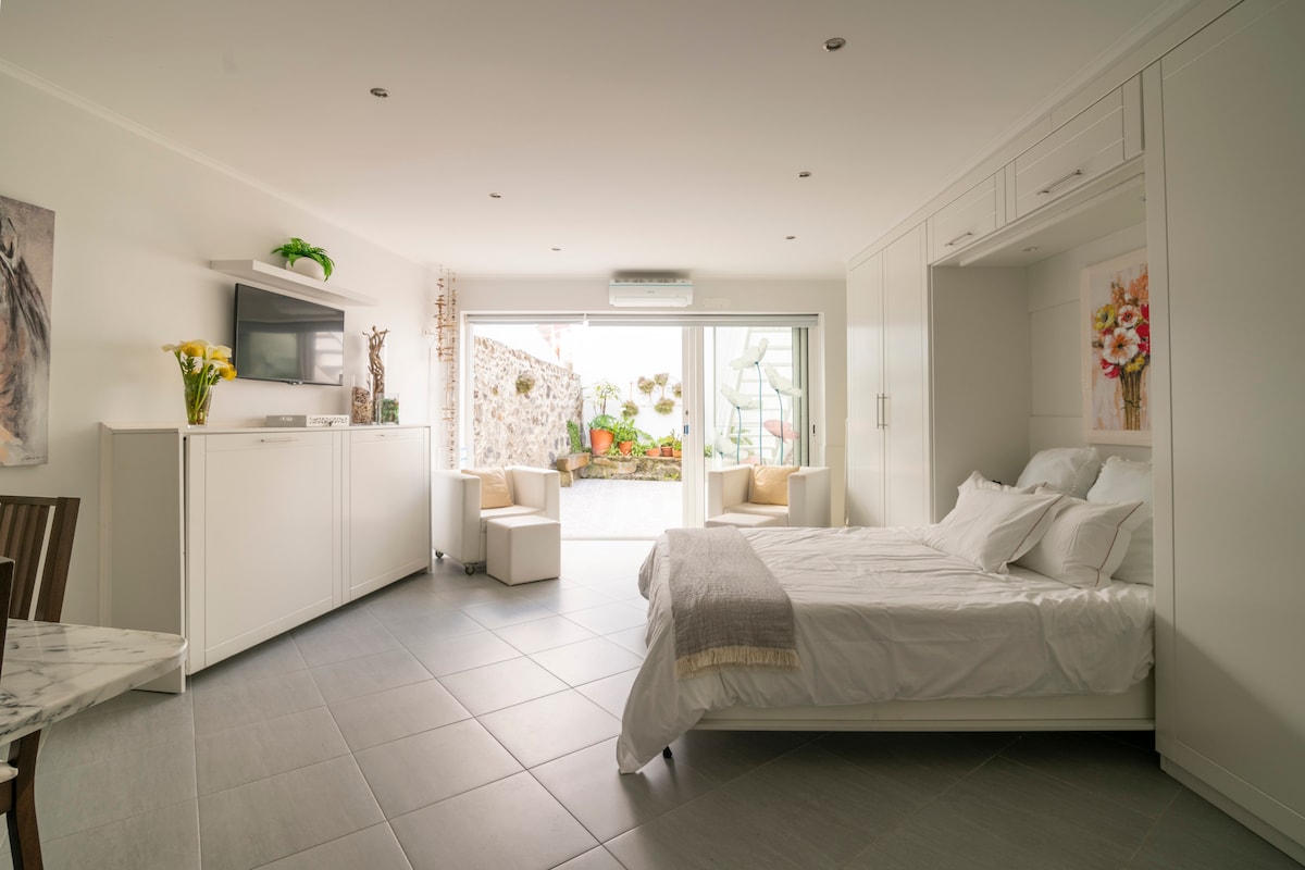 A modern studio space is illuminated by natural light from large sliding glass doors. A spacious bed is neatly made, with a light gray throw blanket draped across it. A small seating area and a television are visible, enhancing the room's functionality.