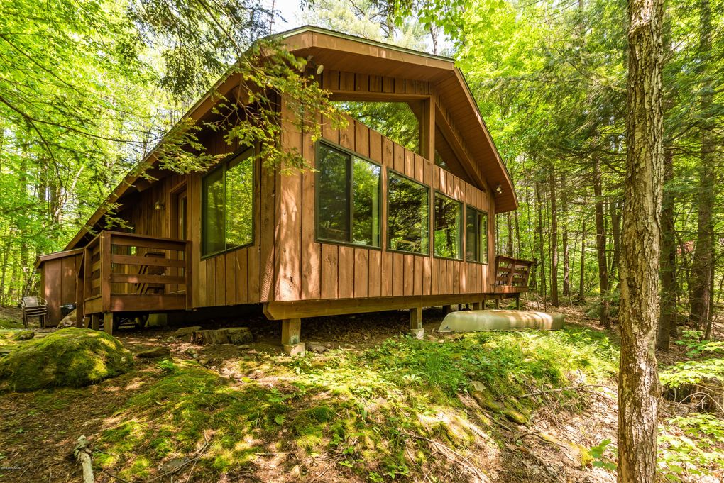 The exterior of the cabin features a sloped roof and large windows, allowing for ample natural light. The wood paneling harmonizes with the surrounding lush greenery. A small deck is visible, offering a seating area to enjoy the serene forest landscape.