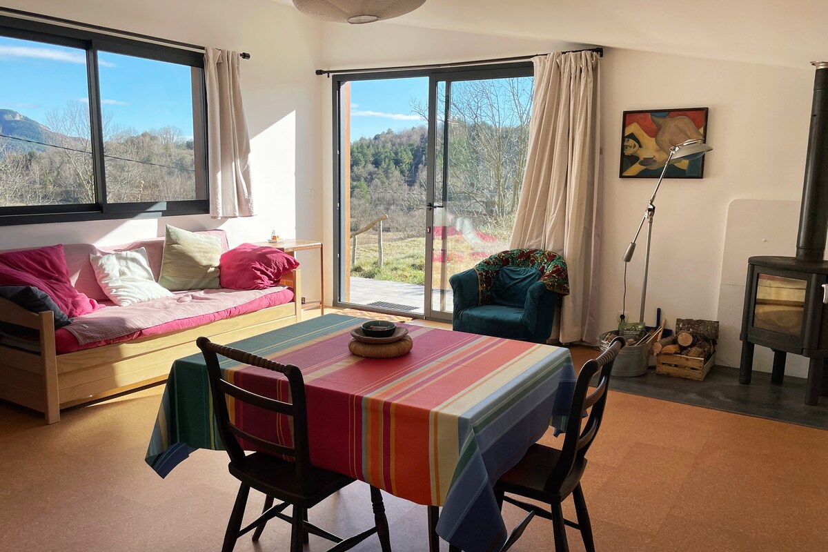 An inviting living space features a colorful striped table set for dining, with chairs positioned around it. A cozy daybed with cushions is situated by large windows, offering views of the surrounding landscape. A wood stove adds warmth to the area.