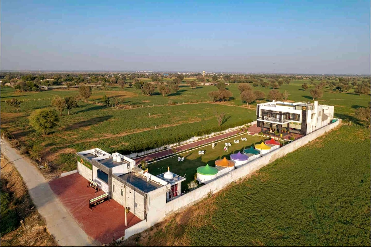 An aerial view of IRIS HEAVEN'Z showcases a spacious farmhouse surrounded by lush green fields. The property features colorful tents arranged in a grassy area, with a modern building positioned alongside a paved driveway. The tranquil landscape emphasizes the connection to nature.