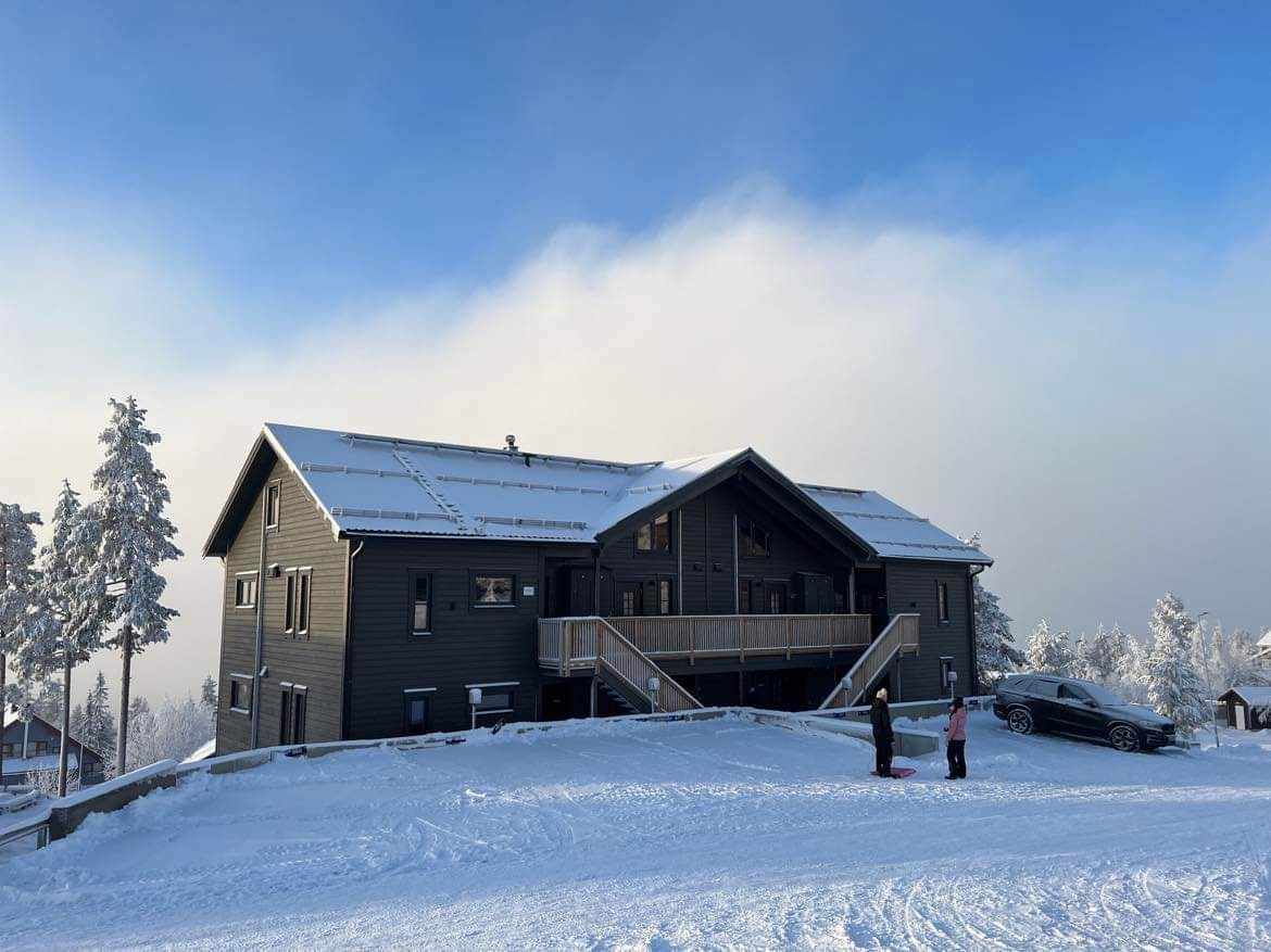 A modern two-story building with a dark exterior is set against a snowy landscape. The roof is lightly dusted with snow, and large deck spaces are visible. Two individuals are seen in the foreground, enjoying the winter scenery, while a car is parked nearby.