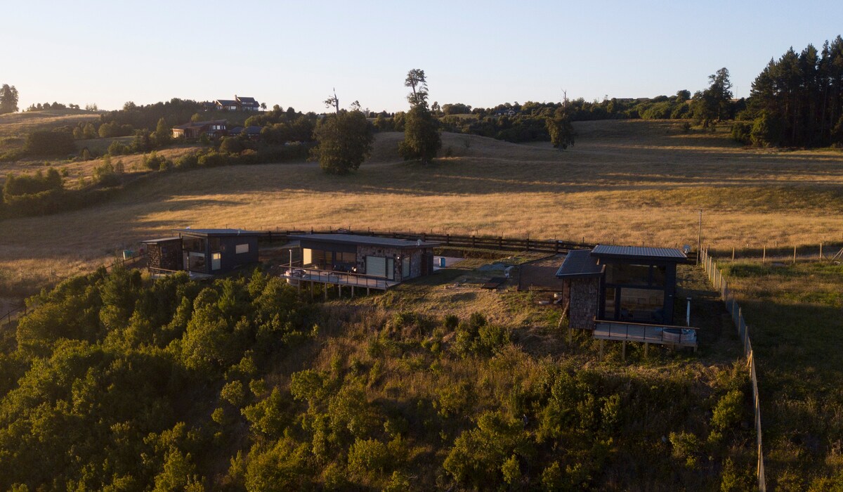 Aerial view of three exclusive cabins nestled in a lush landscape, featuring rolling hills and scattered trees. The cabins are designed with large windows and patios, showcasing their proximity to the natural surroundings and panoramic views of the area.