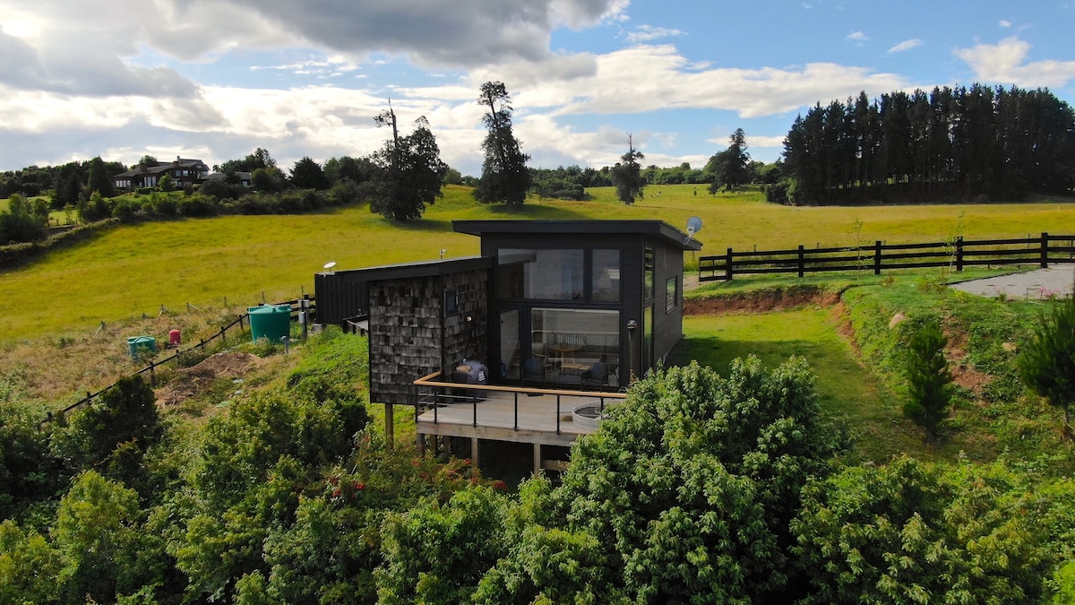 A modern cabin is positioned on a hillside, surrounded by lush greenery. Large windows reflect the sky and offer expansive views of the surrounding landscape. The space features a spacious deck, inviting relaxation while appreciating the natural surroundings and open fields.
