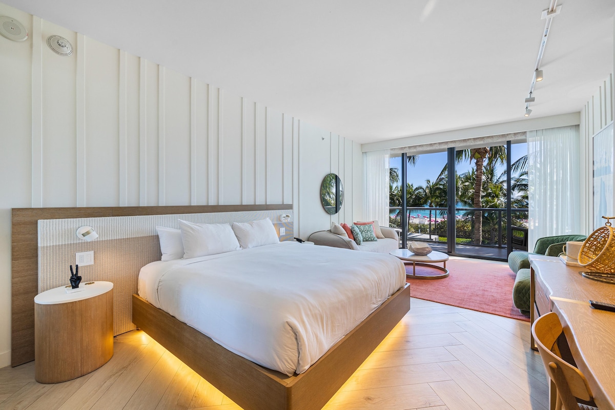 A spacious studio features a large bed with soft bedding, complemented by a comfortable couch and desk area. Floor-to-ceiling windows provide ample natural light and showcase views of palm trees and outdoor spaces. Neutral tones create a calming environment.