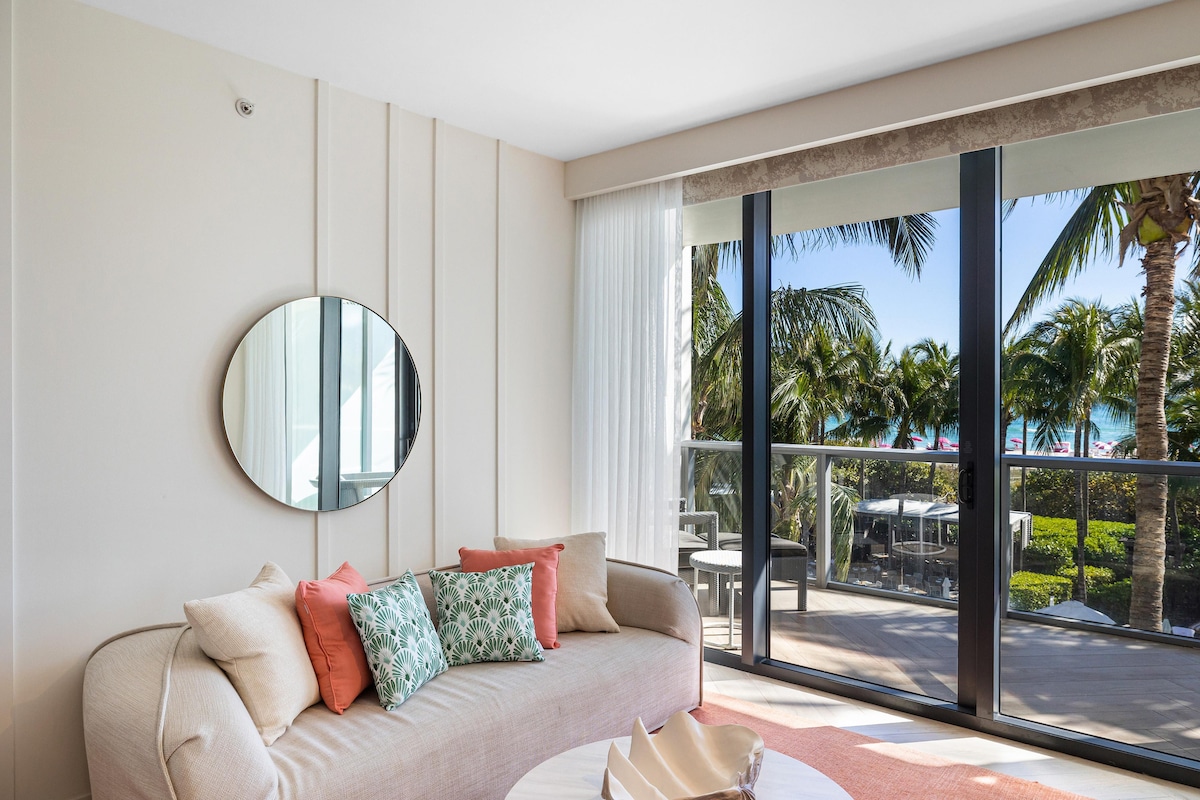 A light-filled living area features a cozy couch adorned with decorative cushions. A round mirror reflects the sunlight, while sliding glass doors provide unobstructed views of palm trees and the ocean beyond. A small balcony table and chairs are visible outside.