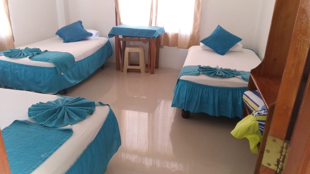 A bright and airy sleeping area is presented with three beds draped in turquoise linens. A small table is positioned between the beds, while natural light filters in through the sheer curtains, illuminating the room's clean and polished floors.