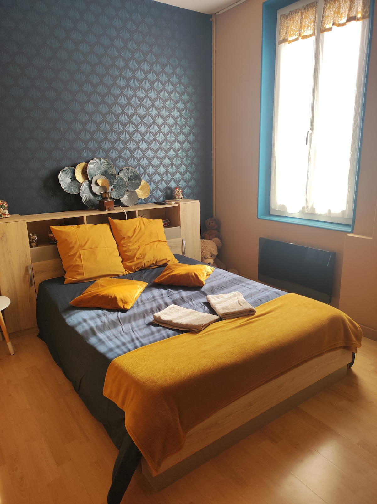 A bright bedroom features a spacious bed adorned with mustard-colored pillows and a coordinating blanket. A wood headboard complements the textured blue wallpaper. Natural light is filtered through a window, illuminating decorative wall elements and a small bedside table.