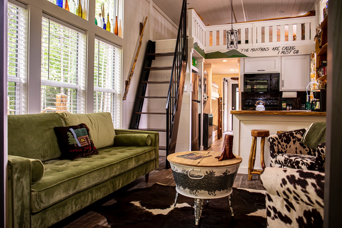 The living area features a green velvet sofa and a cowhide rug, complemented by a rustic wooden coffee table. A spiral staircase leads to an upper loft area. Large windows allow natural light to fill the space, highlighting the cabin's cozy atmosphere.