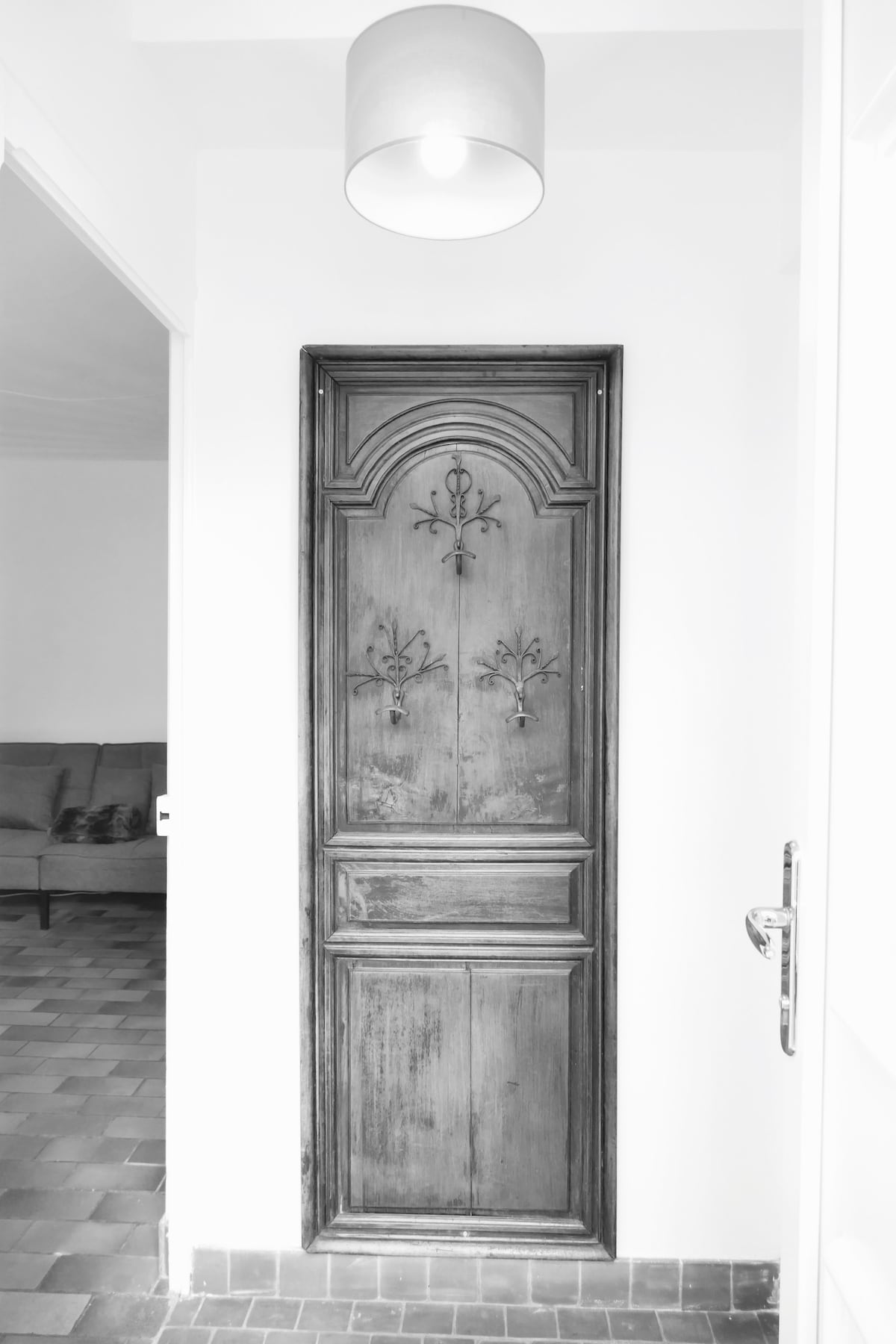 An intricately carved wooden door stands in a bright hallway, showcasing decorative elements etched into its surface. A simple light fixture hangs above, illuminating the space with soft light. The textured floor tiles create a contrast against the smooth walls.