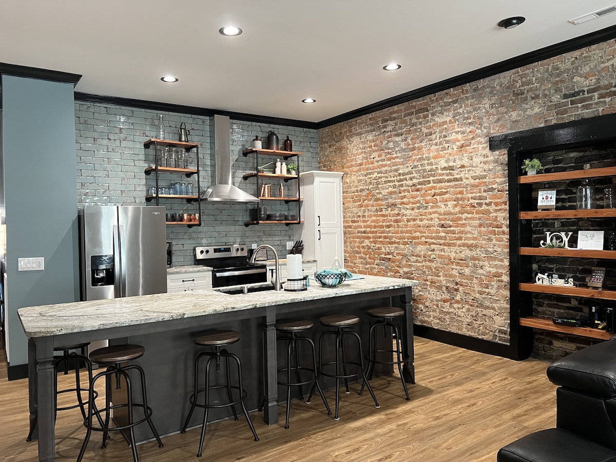 An open kitchen area features a large granite island with seating for four. Exposed brick walls contrast with modern appliances, including a stainless steel refrigerator and a sleek range hood. Wooden shelves display dishware, adding character to the space.
