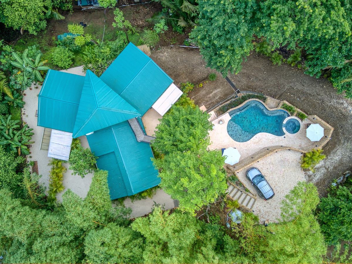 An aerial view showcases the house with a distinctive multi-faceted roof, surrounded by lush greenery. A small pool is visible beside the house, accompanied by sun loungers and umbrellas. A vehicle is parked on a gravel path leading to the entrance, highlighting the tranquil setting.