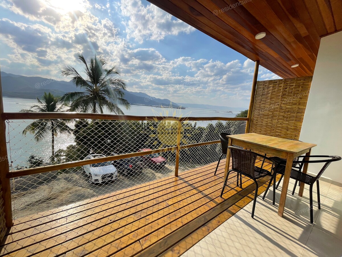A private balcony offers a scenic view of the sea, framed by palm trees. A wooden table and black chairs provide seating, while sunlight illuminates the area. The horizon features distant mountains under a partly cloudy sky.