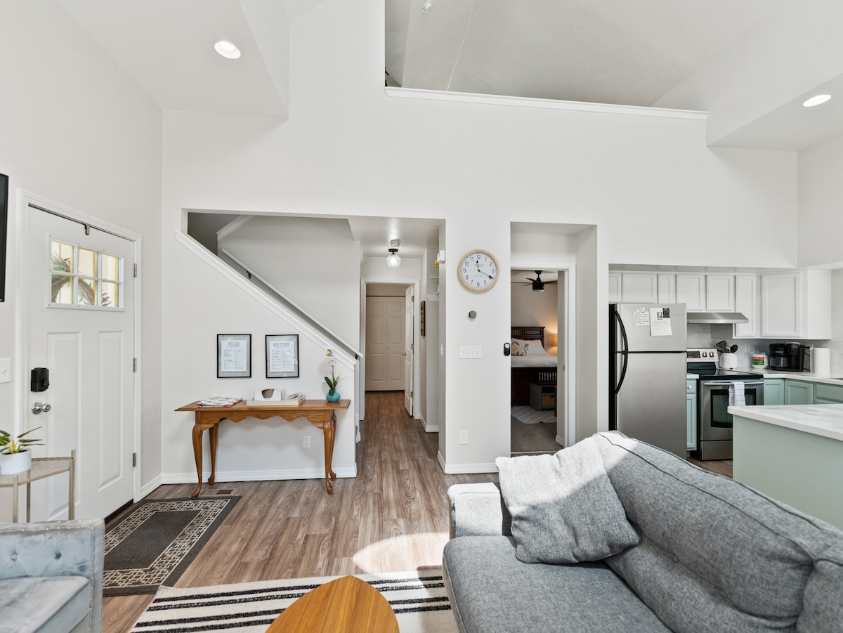 An open living space displays a comfortable gray sofa with armchairs nearby. A wooden table rests against the wall, beside a front door. The view transitions to a kitchen area featuring modern appliances, with a staircase leading to the upper level. Natural light fills the room.