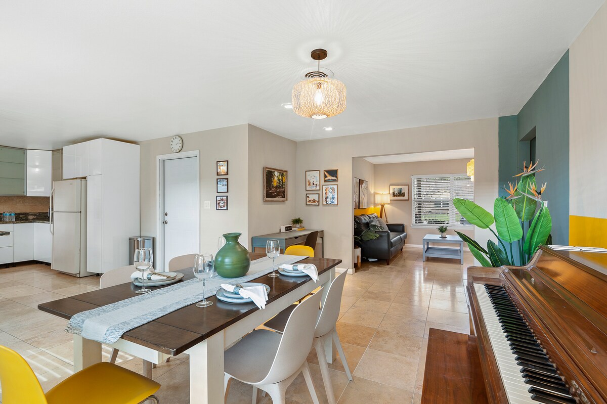 A modern dining area is highlighted by a stylish table set for four, with elegant tableware. A piano is positioned nearby, complementing a spacious living area visible through an archway. Large windows allow for natural light, enhancing the inviting atmosphere.