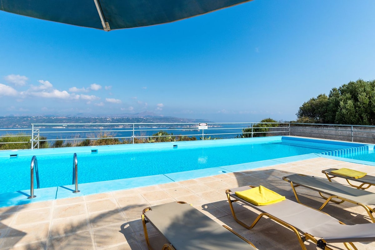 A large swimming pool features a clear blue surface, reflecting the sky. Sun loungers with yellow cushions are arranged around the pool area, providing space for relaxation. The surrounding landscape offers distant views of Souda Bay and lush greenery.