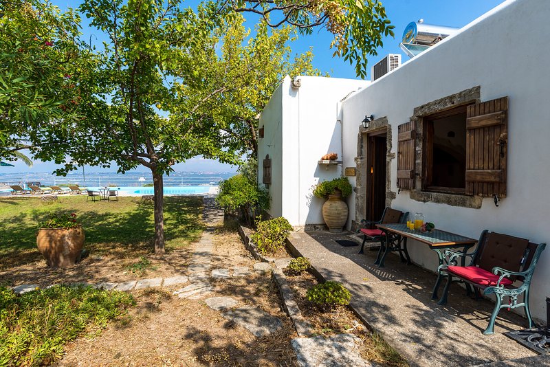 A serene outdoor area is shown, featuring a stone pathway leading to a white cottage. Two chairs with red cushions sit at a small table beneath a tree, with a view of a large swimming pool in the background.