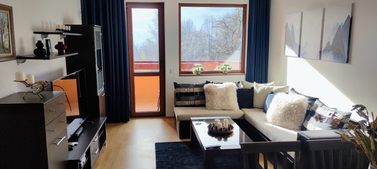 A spacious living area is shown, featuring a comfortable sectional sofa adorned with various cushions. A coffee table sits in the center on a dark rug. Natural light streams through large windows, illuminating the space, while a balcony offers views of the surrounding landscape.