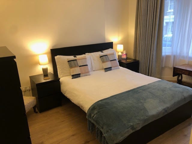 A spacious sleeping area features a large bed with a white quilt and decorative pillows. Soft lighting is provided by bedside lamps on either side, while light-colored curtains frame a window that allows natural light to fill the room.