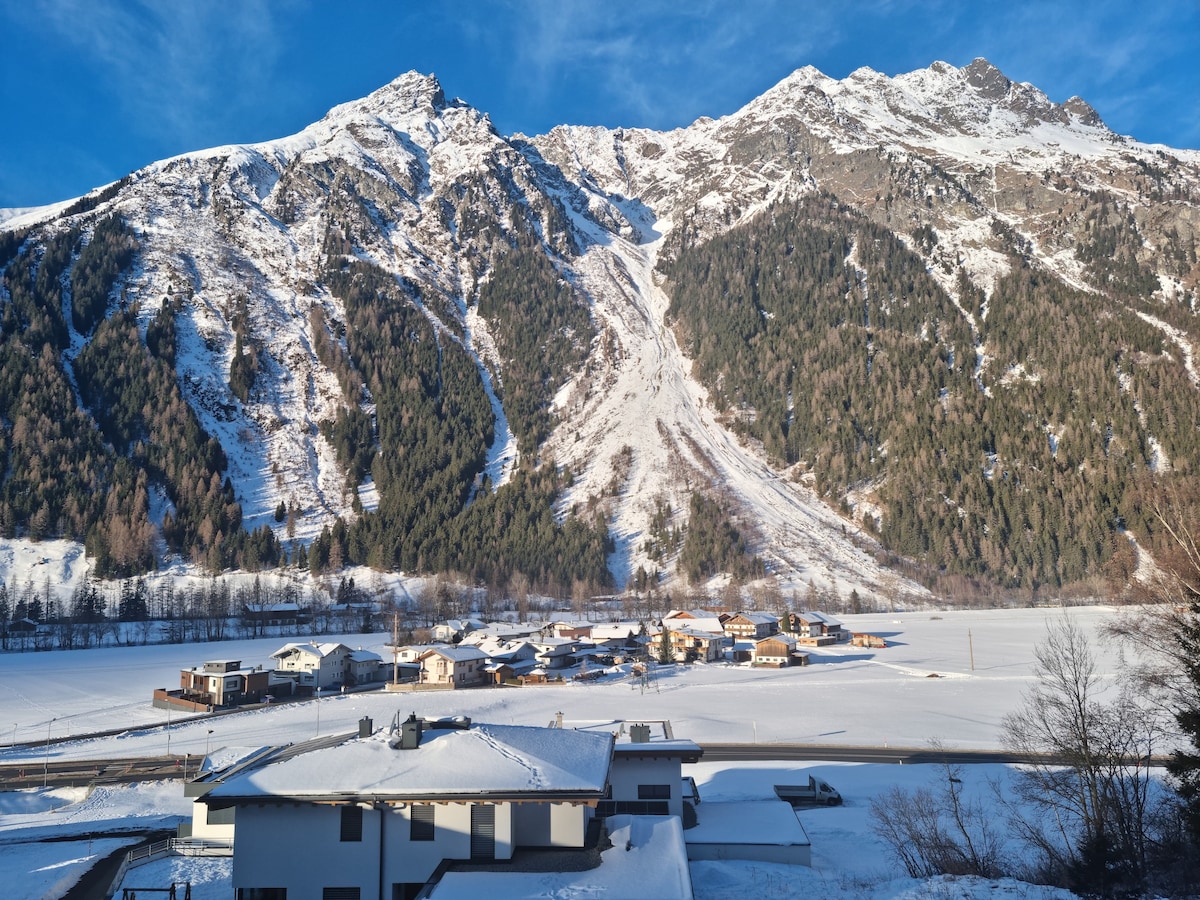 A panoramic view captures snow-covered mountains rising dramatically in the background, with a peaceful village nestled at the foot. The landscape is blanketed in white, while the sun casts a gentle light on the scene, enhancing the serene winter atmosphere.