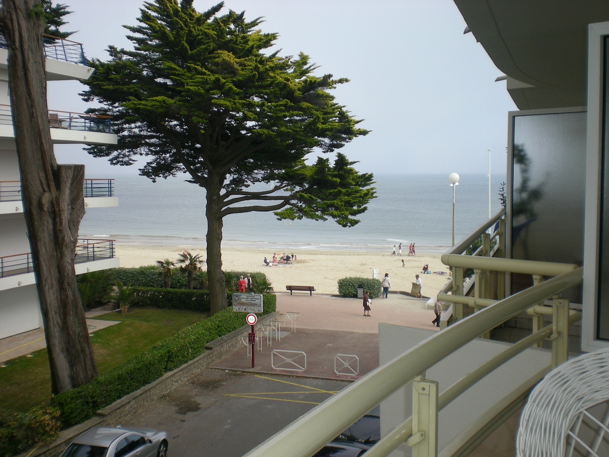 A view from a terrace showcases the sandy beach and ocean beyond, framed by tall trees. Nearby, the pathway is lined with greenery, and individuals can be seen enjoying the beach. The image captures a serene coastal atmosphere.