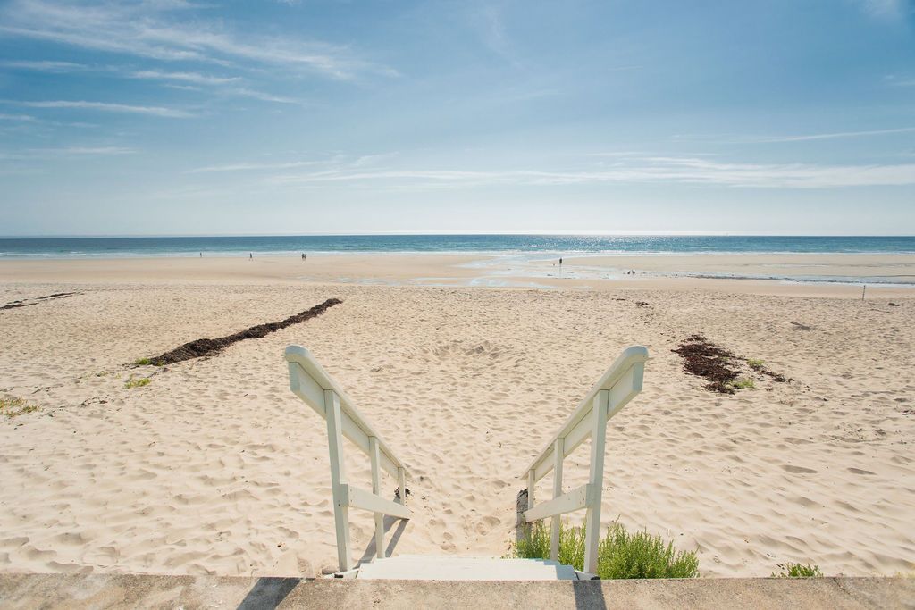 A set of white stairs leads directly from the beachfront property down to the sandy shore. The expansive beach stretches out under a clear blue sky, with gentle waves lapping at the coastline. A few distant figures can be seen enjoying the beach.