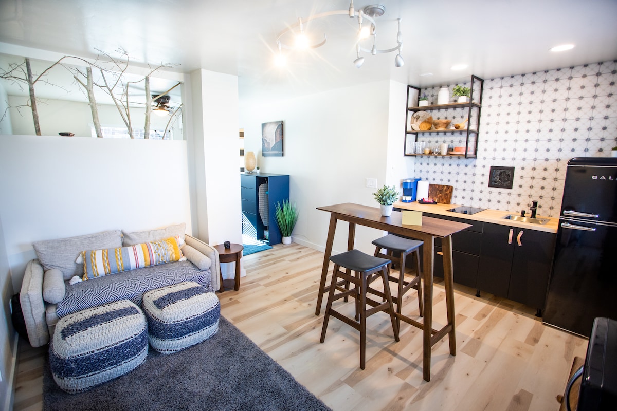 A cozy studio space features a plush couch with colorful striped accents and a small dining area with wooden stools. The modern kitchen, equipped with sleek black cabinetry and a retro-style refrigerator, adds a unique charm. Natural light fills the room, enhancing its inviting atmosphere.