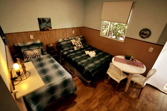 The room features a spacious double bed alongside a single bed, with patterned bedding. A small round table is placed near a window, allowing natural light to fill the space. Simple decor and wooden flooring enhance the warm atmosphere.