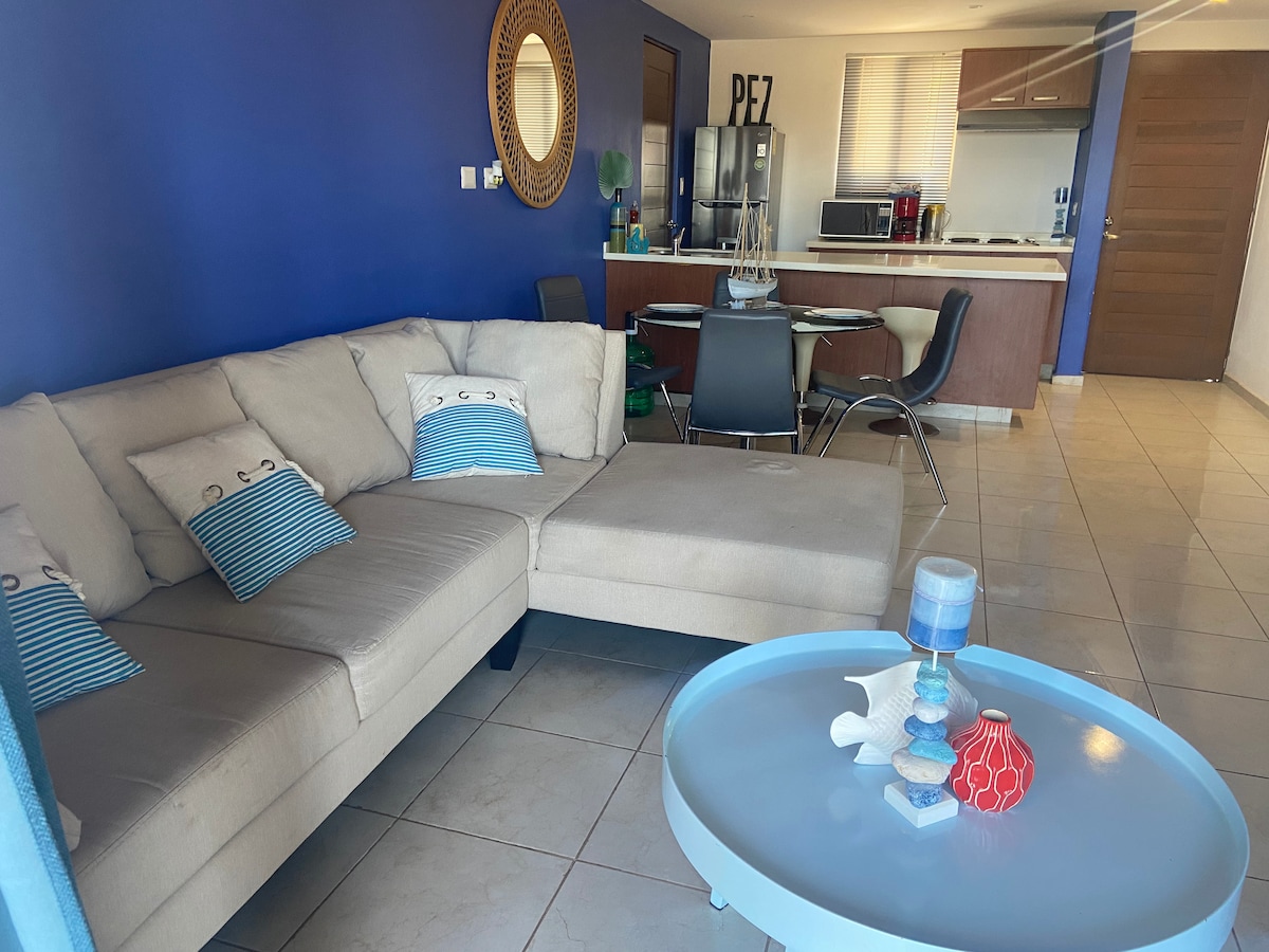 A light-colored sectional sofa is positioned in an open living area, accented with decorative cushions. A round coffee table with various decorative items sits at the center. The kitchen area, featuring modern appliances and wooden cabinetry, is visible in the background.