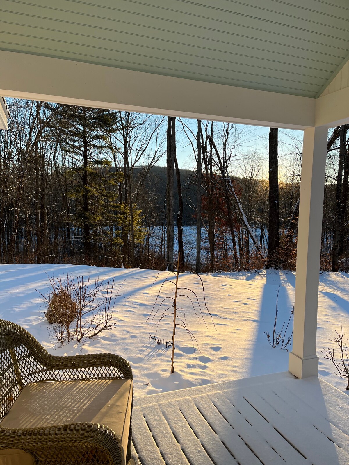A cozy porch area is visible, featuring a wicker chair set against a backdrop of snow-covered ground and trees. Soft sunlight filters through, casting shadows on the porch. A peaceful view of a snow-draped landscape extends towards the horizon.