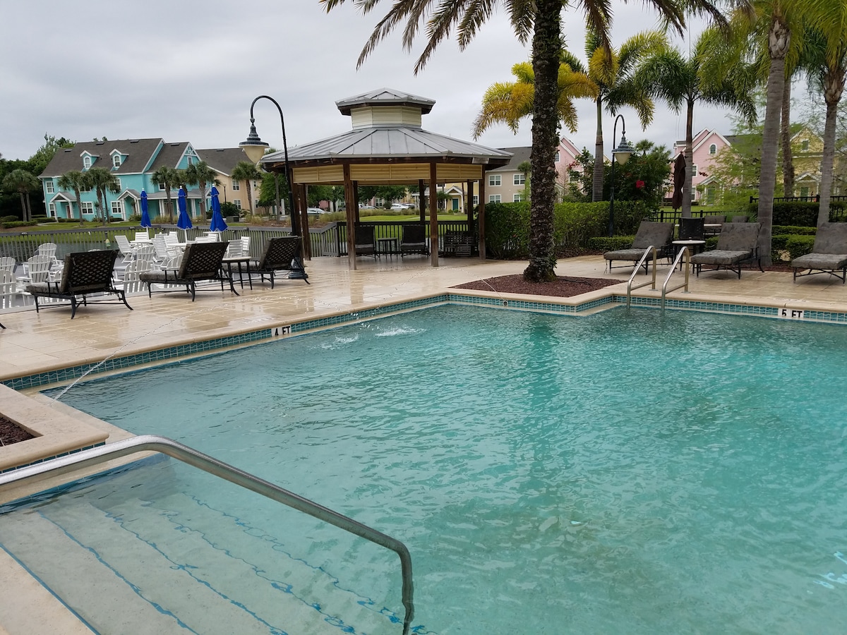 A serene pool area is featured, accompanied by comfortable lounge chairs arranged around the water. Lush palm trees and a nearby gazebo add to the inviting landscape. The pool shows gentle ripples, highlighting a calm atmosphere, while nearby colorful buildings can be seen in the distance.