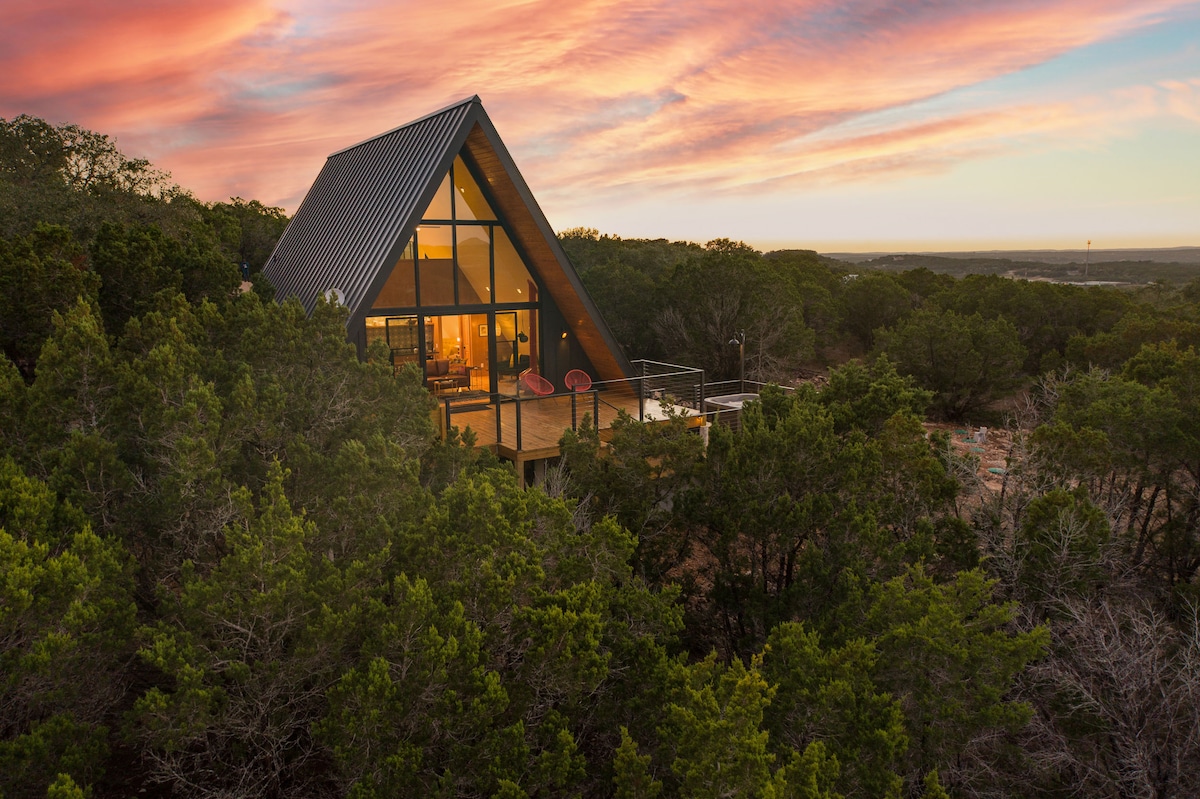An A-frame cabin is nestled among lush greenery, featuring expansive glass windows that reflect the warm hues of a sunset. A spacious deck extends from the cabin, providing outdoor seating with views of the surrounding landscape.