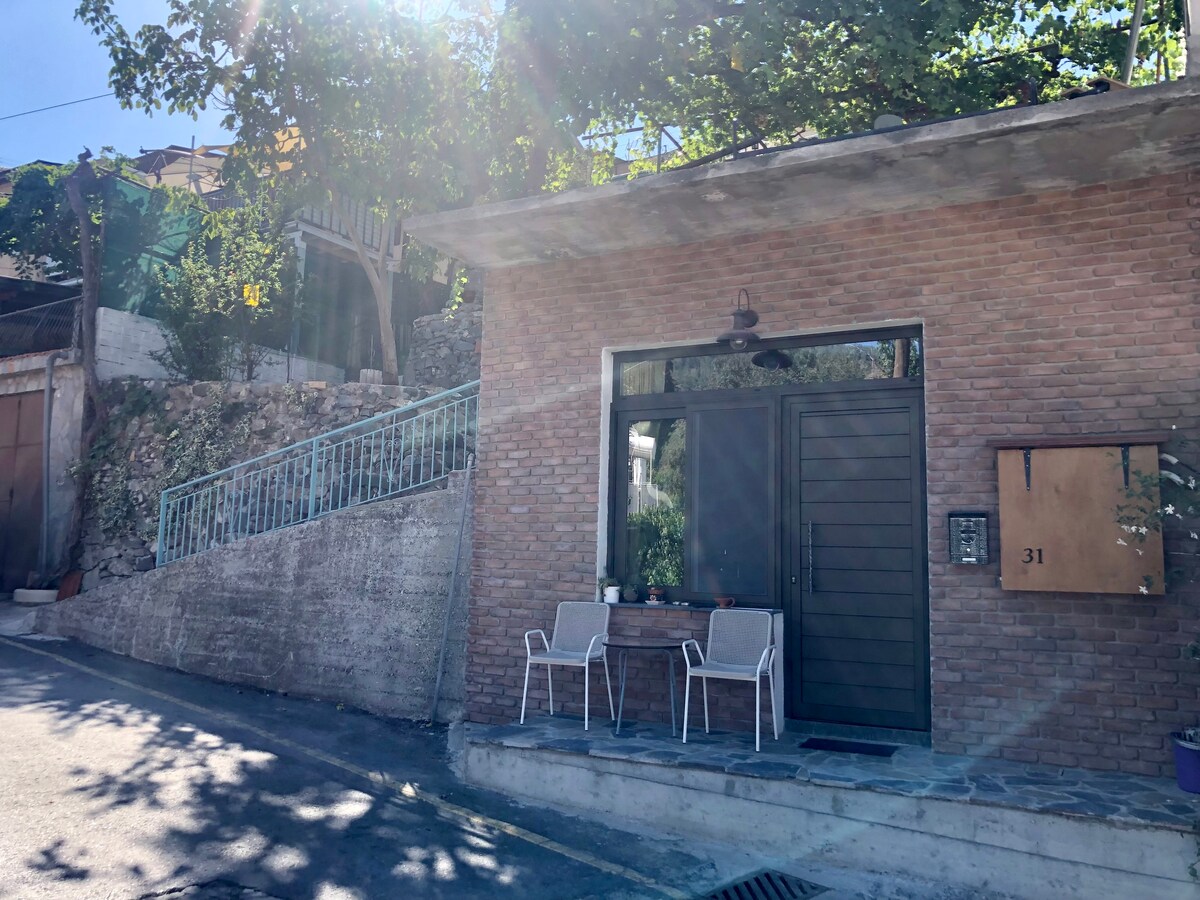 The exterior of the house features a modern brick facade. A wooden door is framed by large glass panels, allowing natural light to enter. Two white chairs and a small table are positioned on the porch, sheltered by greenery from surrounding trees.