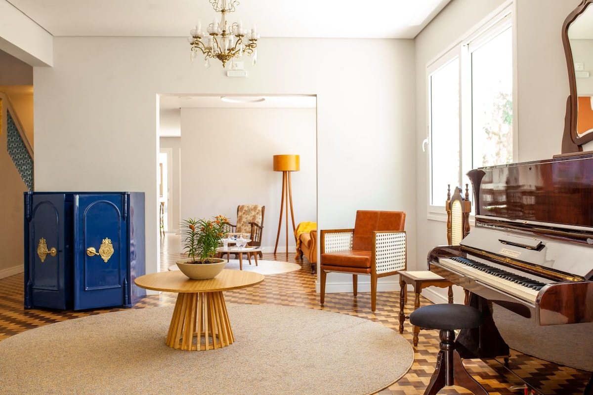 A spacious common area features a wooden piano beside a round coffee table centered on a light-colored area rug. Sunlight enters through large windows, illuminating seating arrangements with a wooden chair, a woven armchair, and a floor lamp in the background.