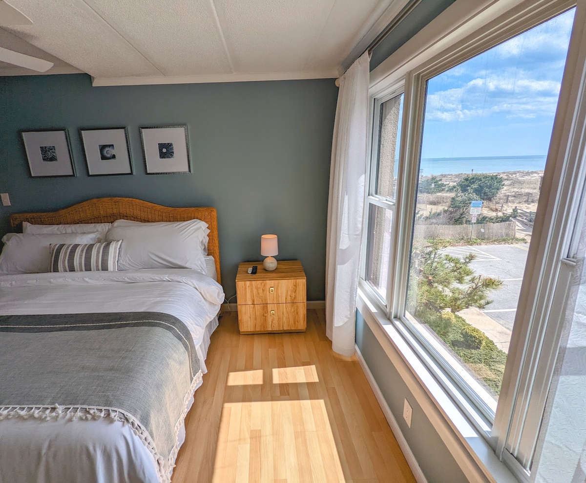 A serene bedroom setting features a spacious bed adorned with crisp white linens, positioned beside a wooden nightstand. Natural light streams in through large windows, offering a view of sandy terrain and the distant ocean. Soft blue walls enhance the tranquil atmosphere.