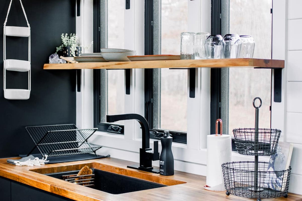 A modern kitchen space features a wooden countertop with a built-in sink. Above, open shelving displays plates, glassware, and decorative items. Large windows provide natural light, while a sleek black faucet and dish rack enhance functionality. A black wire basket and a white dish dispenser are also visible.