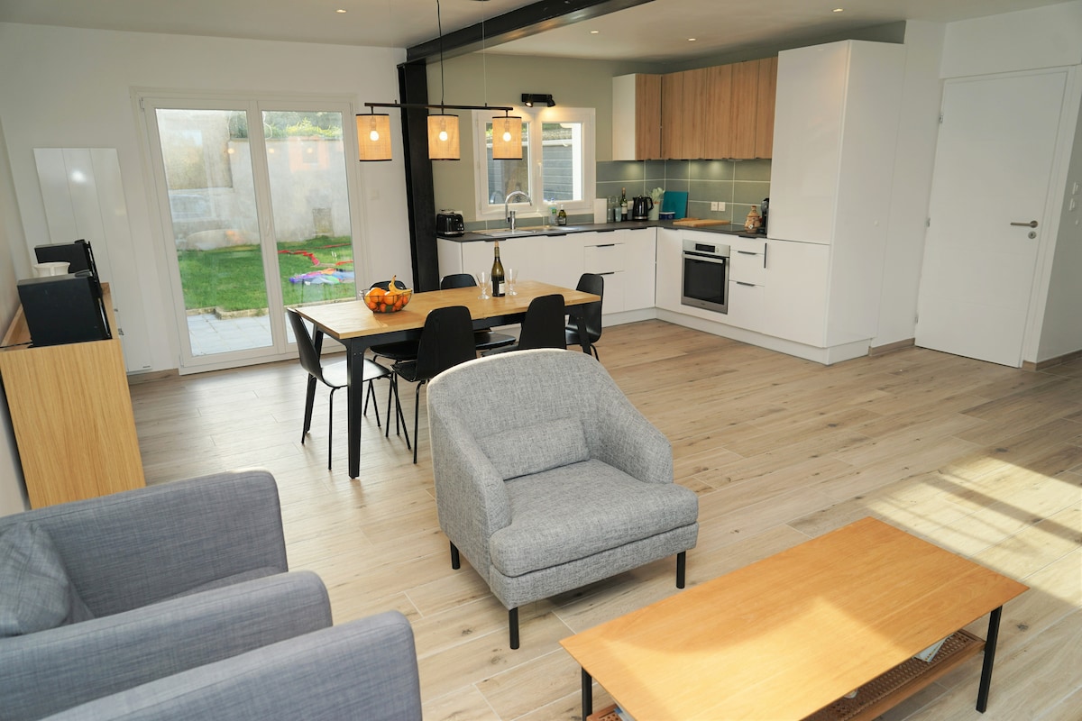 A spacious open-plan living area combines a modern kitchen and dining space. The kitchen features contemporary cabinetry and equipped appliances, while a dining table for six, extendable to ten, is positioned nearby. Two comfortable chairs are set before a wooden coffee table, with natural light streaming through glass doors.