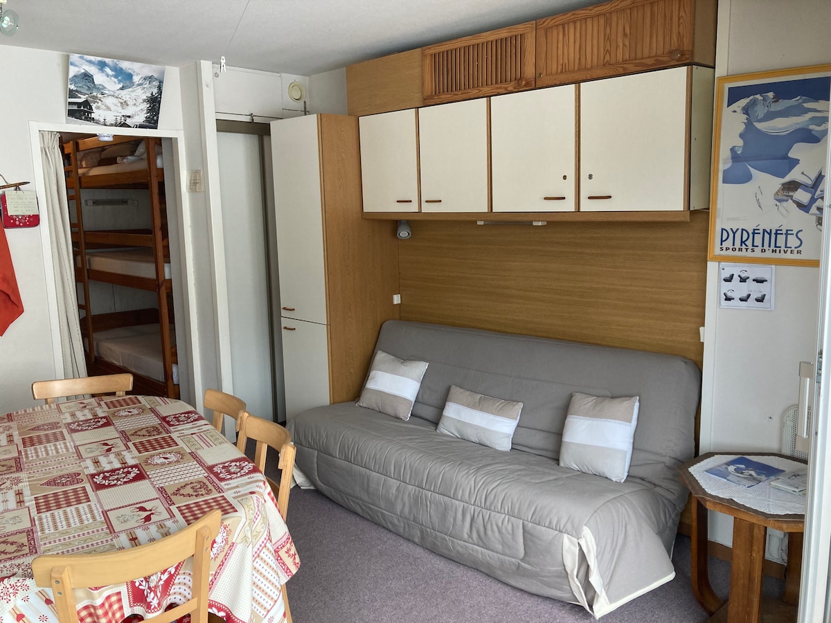 A lounge area is visible, featuring a grey sofa bed against a wooden wall unit with cabinets above. A dining table with light-colored chairs occupies the space, while a framed poster and wall art add interest. A glimpse of a bunk bed can be seen through an open door.