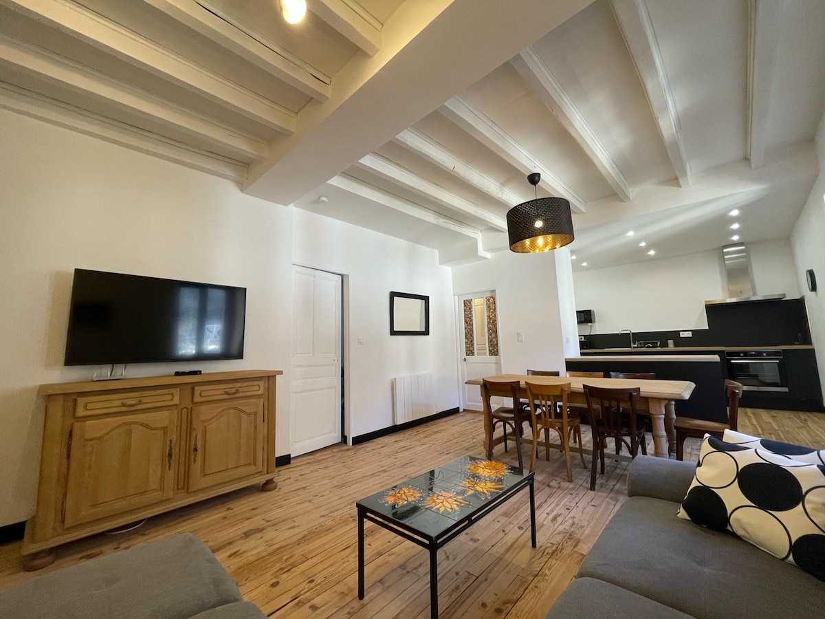 A spacious living area is presented with wooden flooring and high ceilings. A large television is mounted on the wall, while a comfortable sofa and a dining table accommodate several guests. Light fixtures create a warm ambiance, complementing the neutral wall tones.