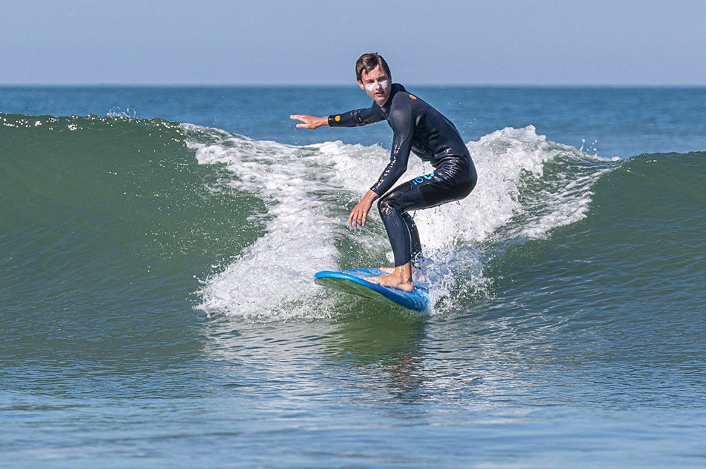 A surfer is seen riding a wave on a sunny day. The ocean stretches out in the background, with gentle rolling waves and a clear sky. The surfer is positioned on a vibrant blue surfboard, demonstrating balance and skill as water splashes around.