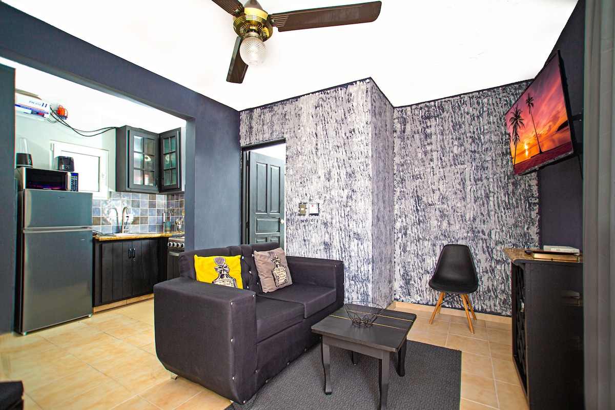 A stylish living area is presented with a black couch and a small table, complemented by a sleek chair. A television mounted on the wall shows a scenic image. The adjacent kitchen area is visible, featuring dark cabinetry and appliances, with blue tile accents.