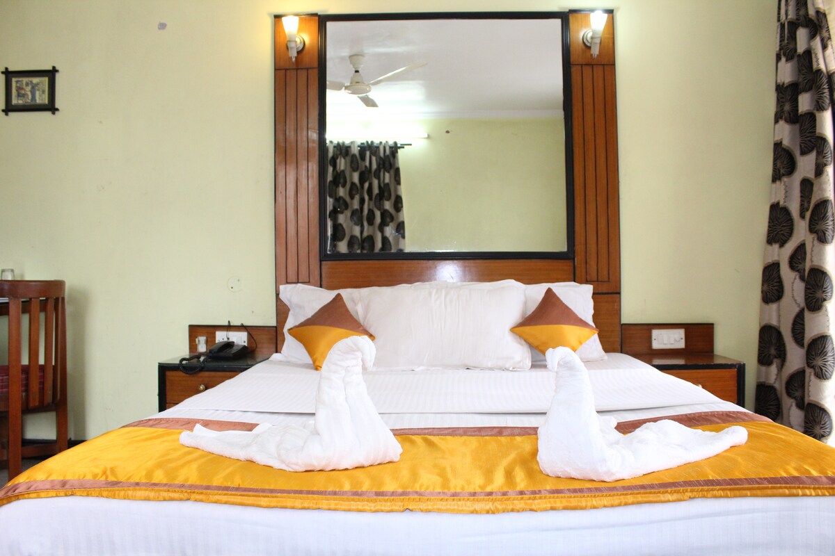 A spacious bed with white linens is centrally located in the room, accented by two decorative pillows. Towel sculptures are arranged on the bed. A large mirror reflects the interior, accompanied by a dark wood nightstand and a chair positioned nearby.