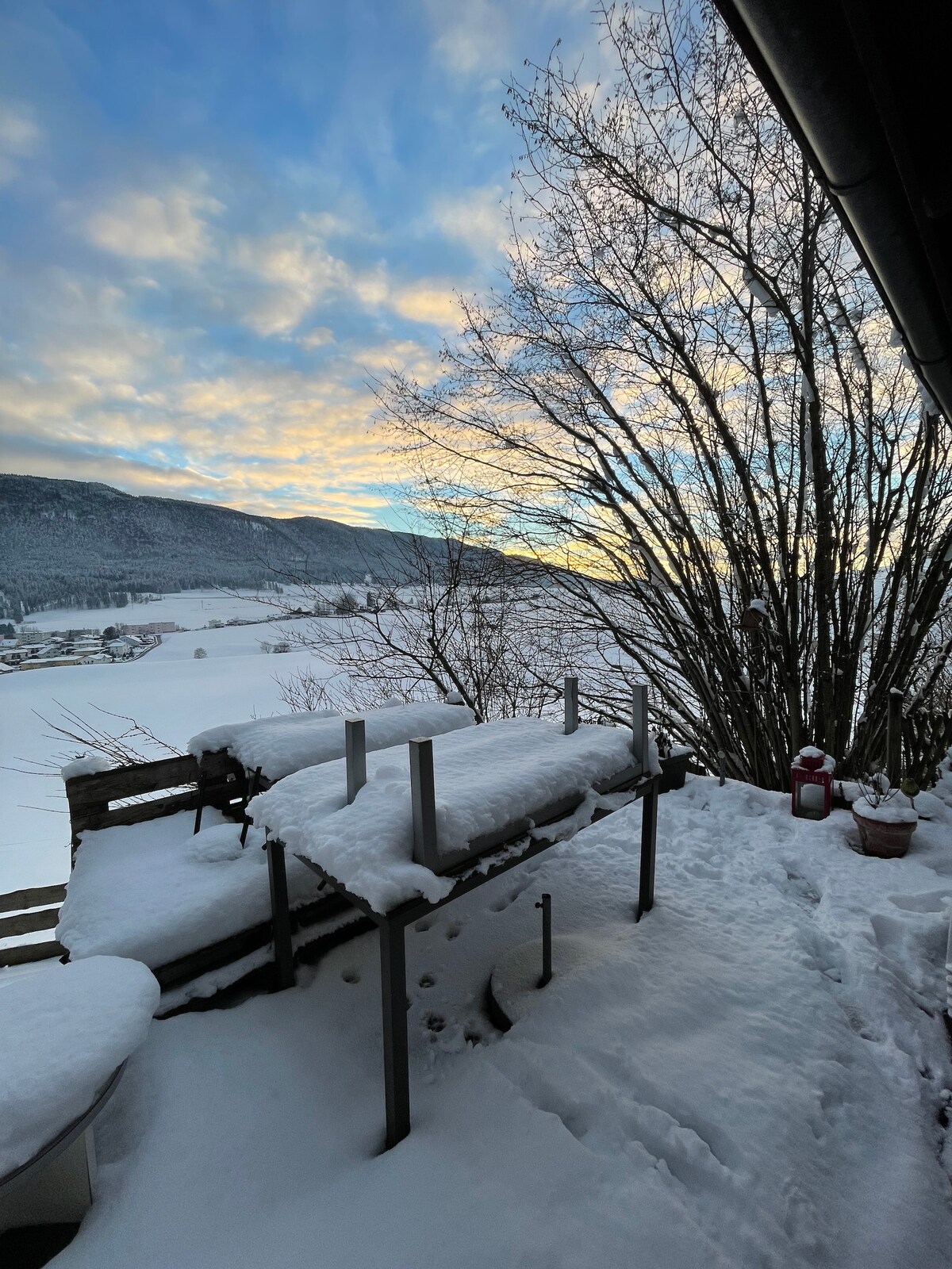 A snow-covered outdoor space is visible, featuring a wooden deck with seating and a railing. The surrounding landscape includes snow-blanketed fields and distant mountains, partially illuminated by the soft light of a cloudy sky. A tree is seen nearby, adding to the winter scene.