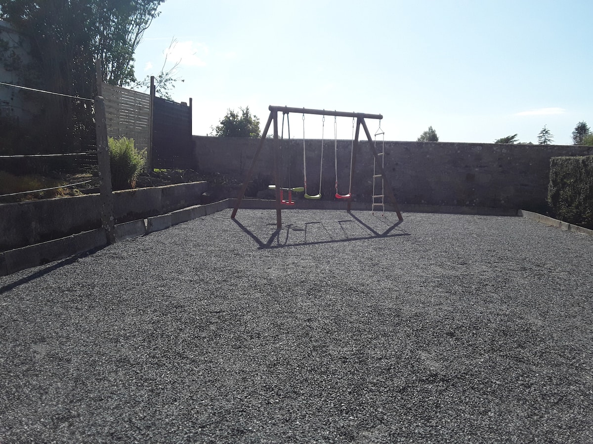 An outdoor space features a gravel surface with a wooden swing set positioned in the center. Surrounding areas are lined with low stone walls and greenery, providing a secure environment for family activities.