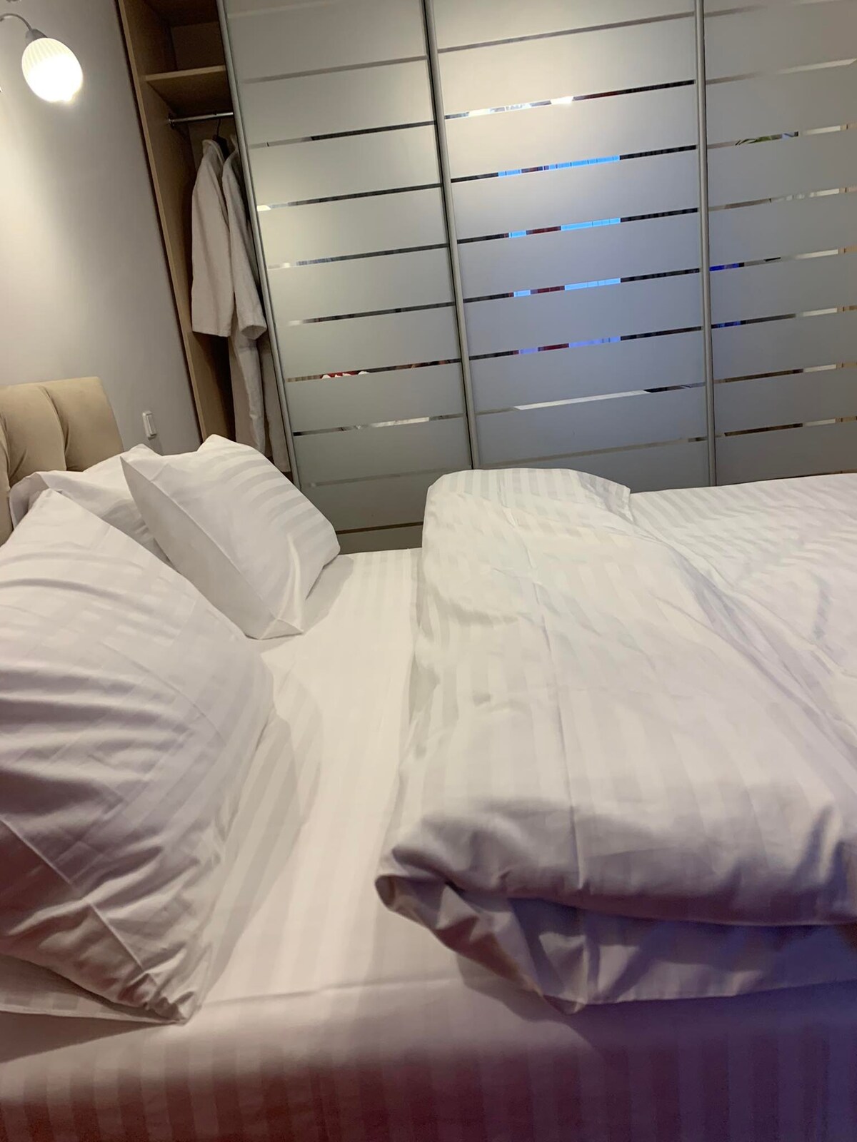 A spacious bed is positioned with neatly arranged pillows. A soft, light-colored blanket is placed at the foot of the bed. Behind it, modern sliding glass doors introduce a subtle design element, while the overall color scheme remains neutral.