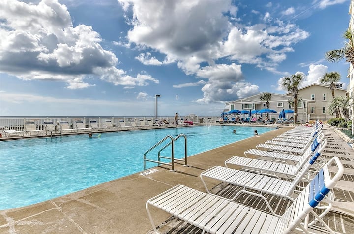 A spacious swimming pool is surrounded by lounge chairs and offers views of the ocean in the background. The pool area is partially shaded by the clouds, while guests can be seen enjoying the water. Palm trees line the poolside, enhancing the coastal ambiance.