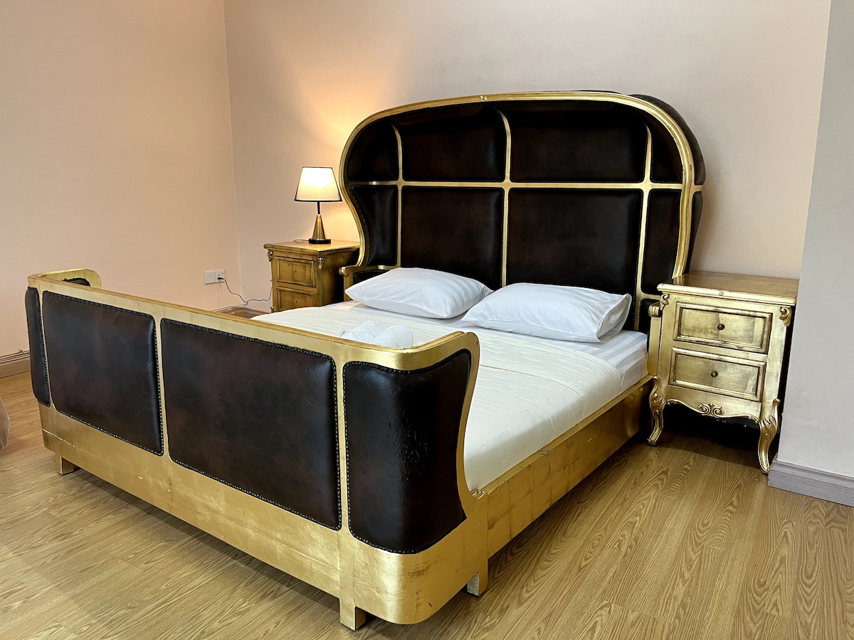 A spacious bed with a distinctive golden and dark wooden headboard is present, accompanied by two bedside tables, each with a lamp. Soft bedding is arranged neatly, and natural light brightens the room, highlighting the warm wooden flooring.