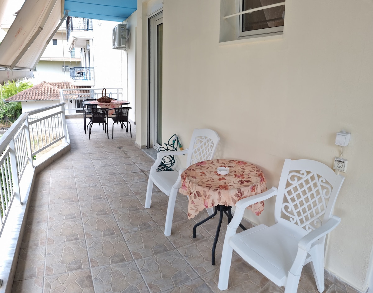 An outdoor balcony features a round table with a floral tablecloth and two white chairs. The space is accompanied by additional seating, and tiled flooring extends across the area. Nearby, views of surrounding buildings and greenery are visible between the railings.